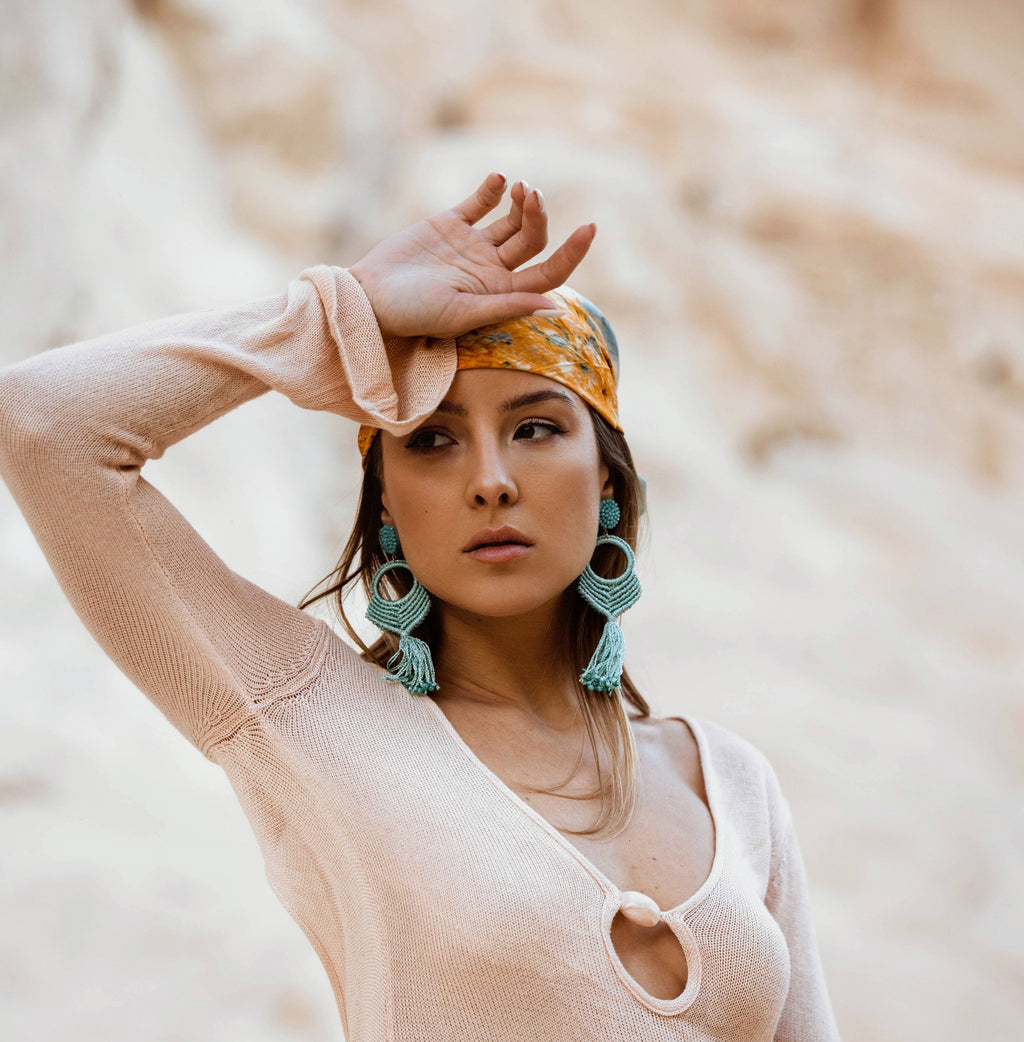 A woman posing outdoors in a light beige knit long-sleeve dress with a keyhole cutout, accessorized with a yellow patterned headscarf and large turquoise earrings.