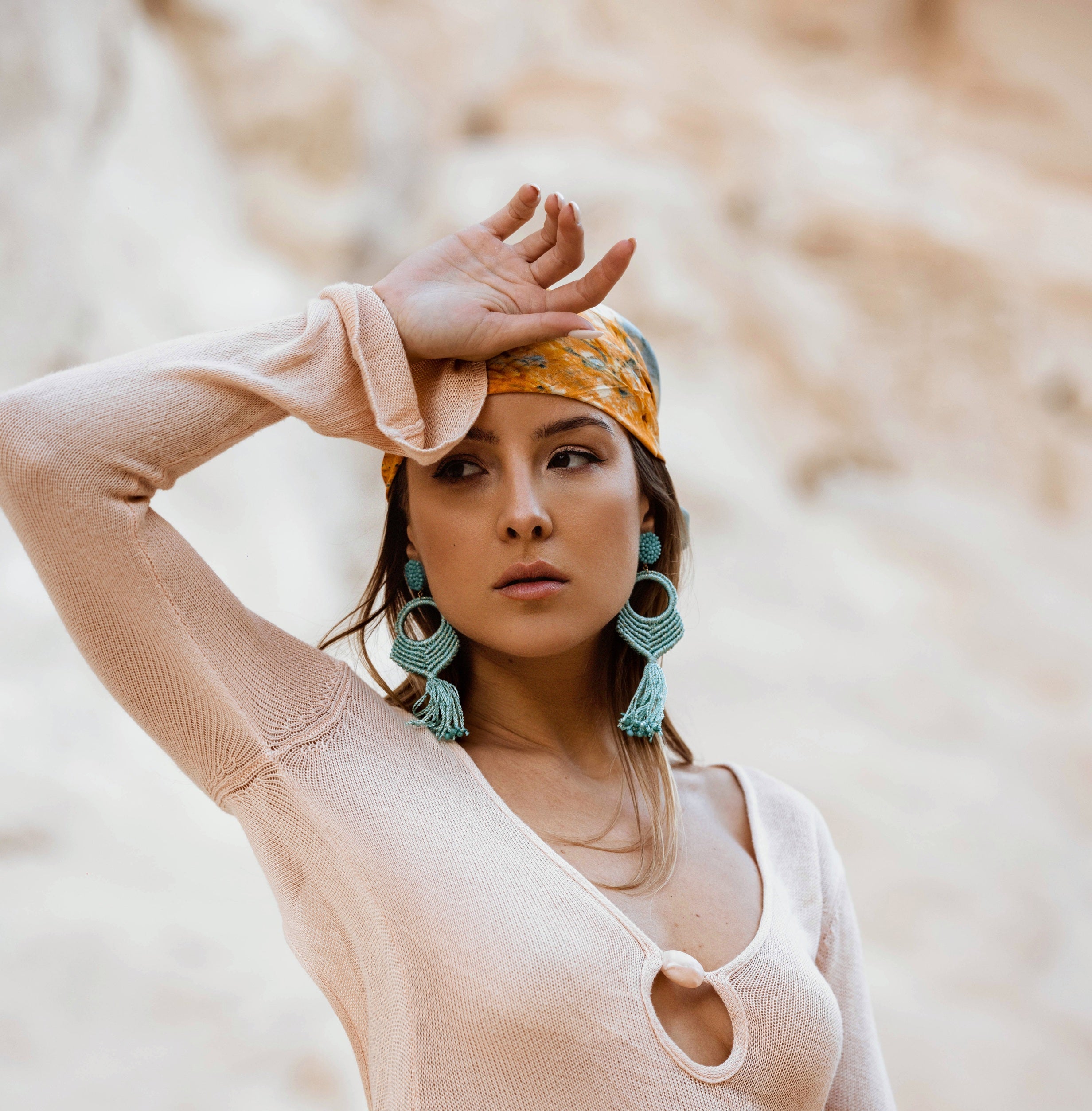 A woman posing outdoors in a light beige knit long-sleeve dress with a keyhole cutout, accessorized with a yellow patterned headscarf and large turquoise earrings.