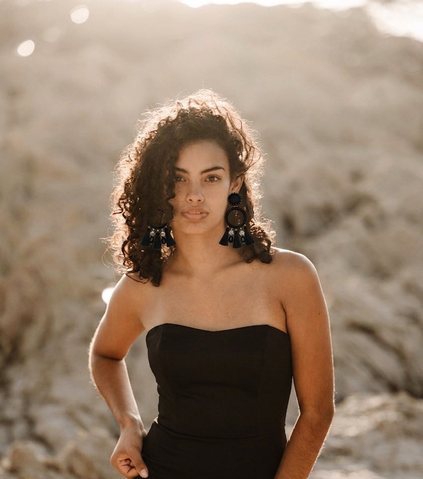 A woman with curly dark hair stands outdoors against a rocky, sunlit background wearing a strapless black dress and large dark statement earrings.