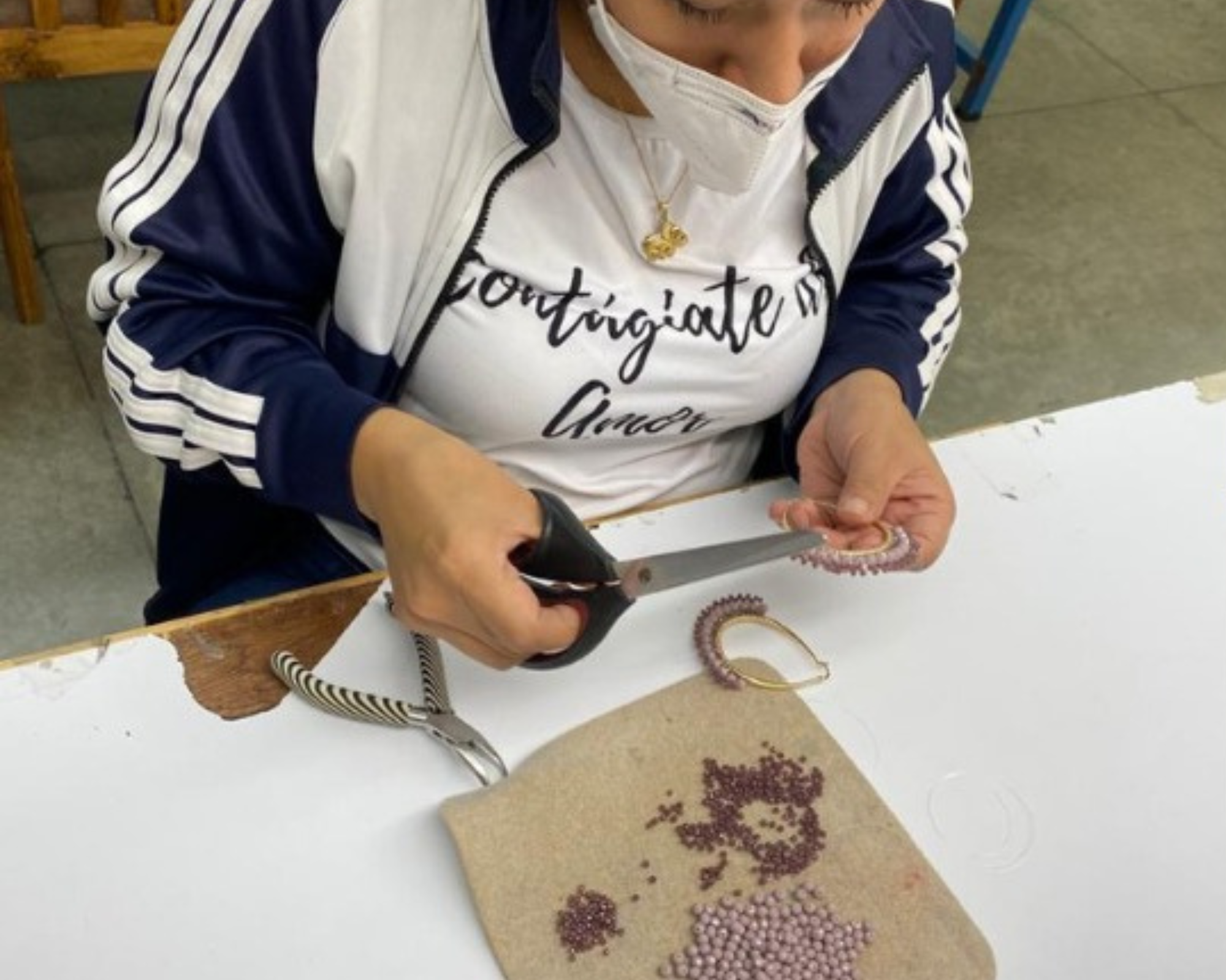 A person wearing a navy and white track jacket and a white face mask sits at a white table, using scissors to cut a strand of small purple beads. A finished beaded bracelet and a fabric pad with bead remnants are on the table.