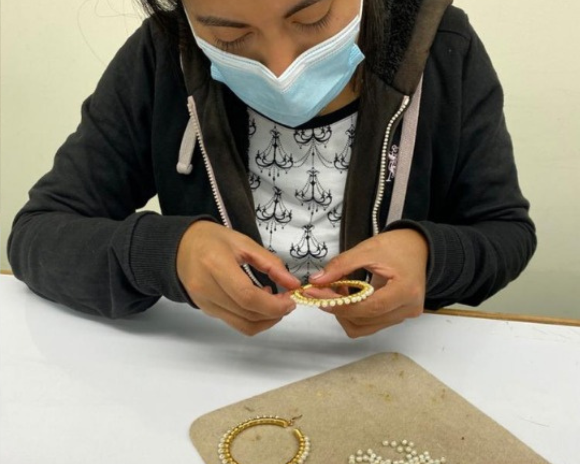 A person wearing a light blue face mask and a black zip-up hoodie sits at a white table, holding a small gold bangle decorated with white pearls. Another pearl bangle rests on a beige mat on the table.