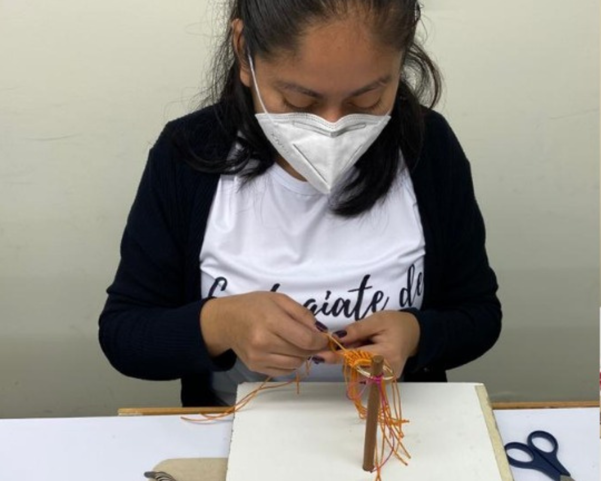 A woman wearing a white face mask and a white t-shirt with a dark navy cardigan is shown from the chest up, sitting at a table and weaving a small orange fiber art piece on a wooden loom.