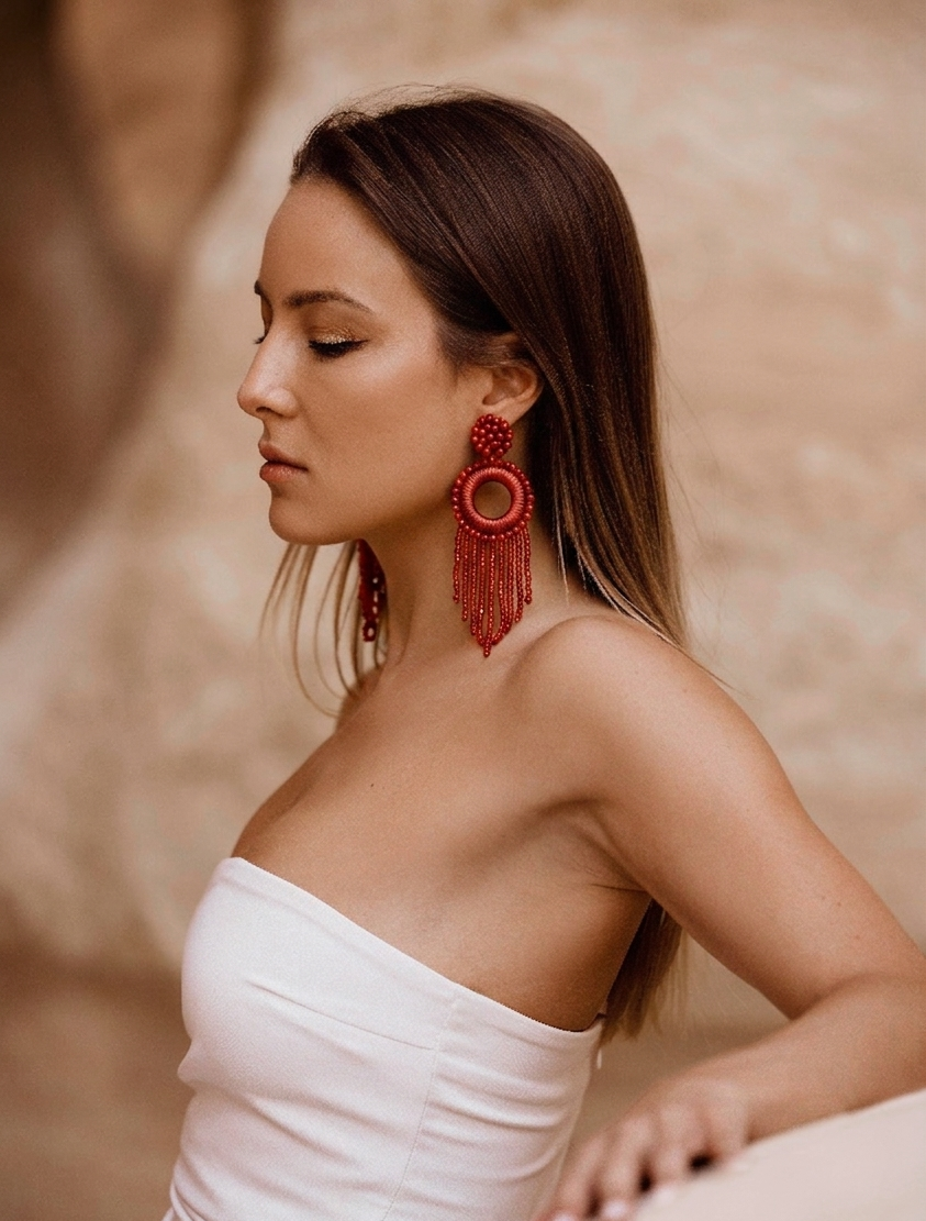 A woman in a white strapless top wearing large red beaded fringe earrings, posed against a warm beige textured background.