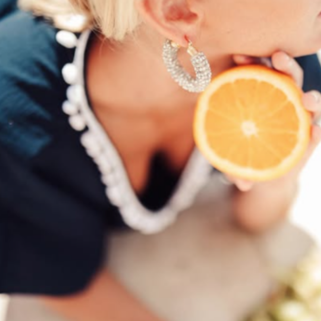 A close-up lifestyle shot of a woman wearing large, sparkling silver hoop earrings while holding an orange slice near her mouth.