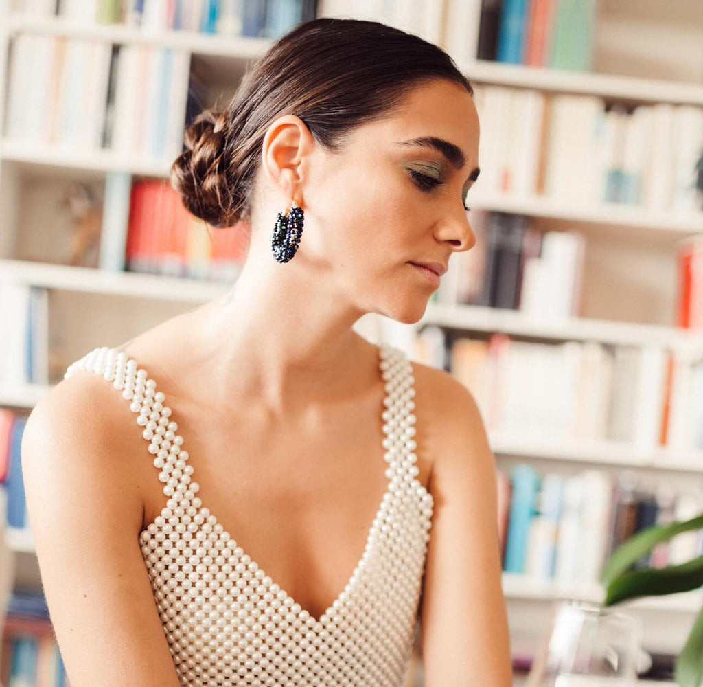 A woman wearing a pearl-beaded sleeveless top and black beaded statement earrings, seated in front of a blurred bookshelf background.
